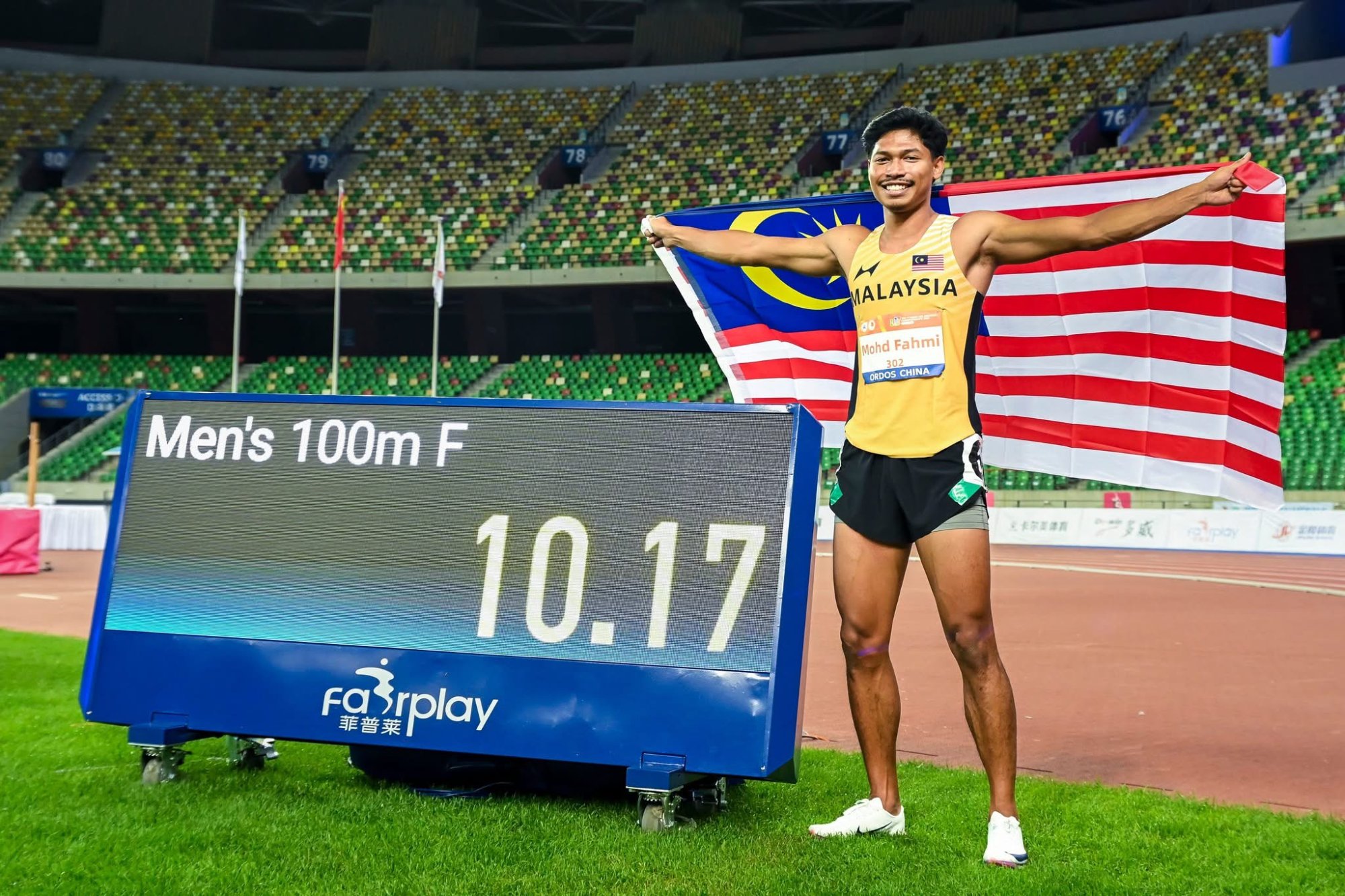 Azeem Fahmi holding the Malaysian flag beside a men’s 100m final results board