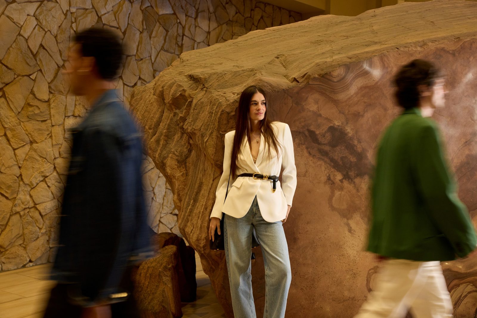 Stylish guest in a white blazer posing against a sculpted sandstone feature as people walk past